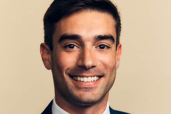 A front on profile of a man wearing a full suit with a red tie, white shirt and a blue jacket with a white pocket square. The man has short brown hair that has been put into a quiff. He is smiling with his teeth and standing in front of a peachy coloured background