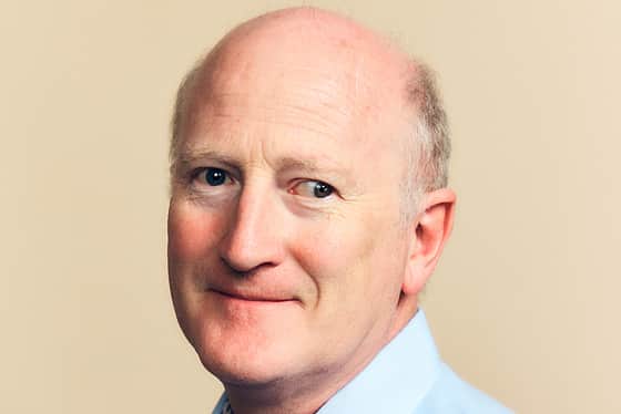 A side on profile of a man wearing a light blue shirt. The man has grey hair either side of his head. He is smiling and standing in front of a peachy coloured background