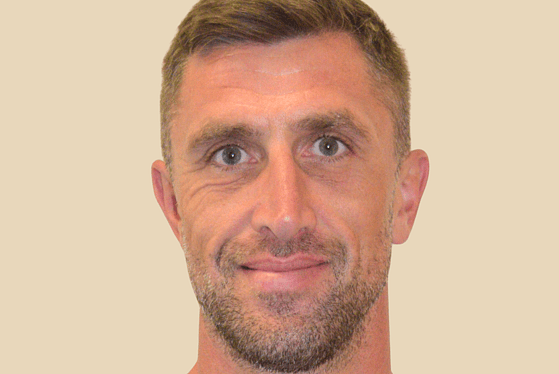A front on profile of a man wearing a navy t-shirt with the Adelphi logo embroidered in on white. The man has short brown hair that has been put into a quiff. He is smiling and standing in front of a peachy coloured background