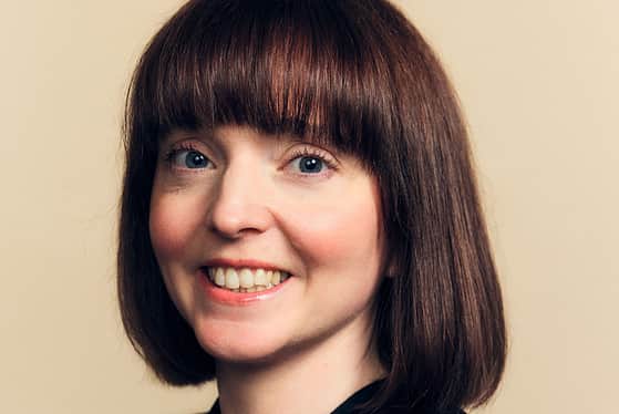 A side on profile of a female wearing a black top. The female has a red bob. She is smiling with her teeth and standing in front of a peachy coloured background