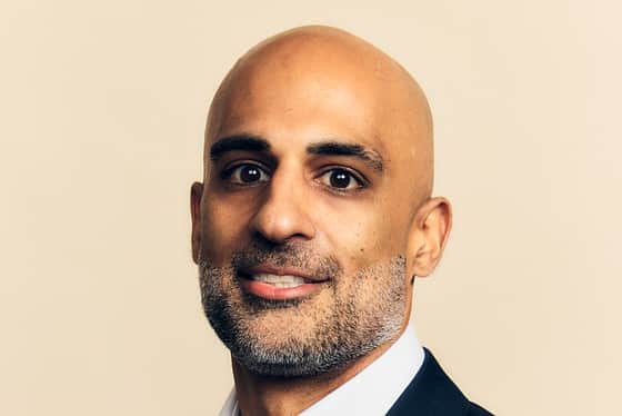 A side profile of a man wearing a full suit with a white shirt and a black jacket. The man is bald and he is smiling with his teeth, standing in front of a peachy coloured background