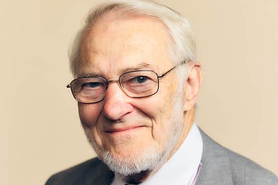 A side on profile of a man wearing a full suit with a turquoise tie, white and red stripy shirt with a grey jacket. The man has short white hair that has been swept back. He is smiling and standing in front of a peachy coloured background
