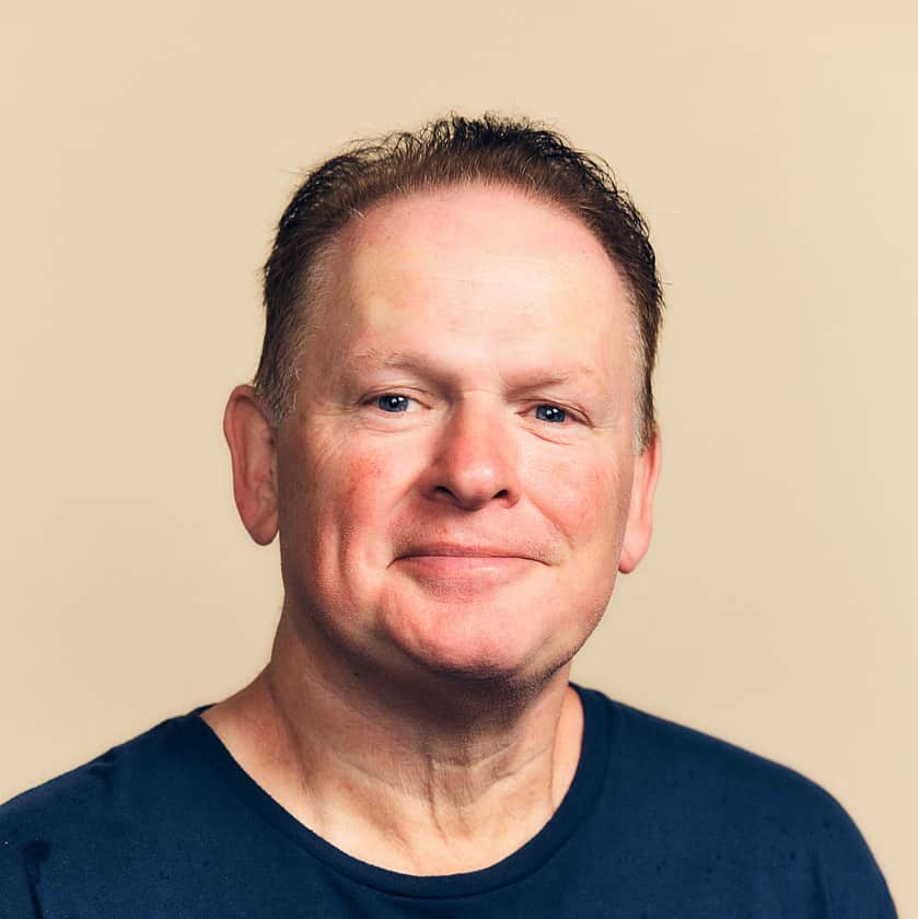 A smiling man with brown hair, wearing a navy blue t-shirt with an Adelphi logo on the chest.