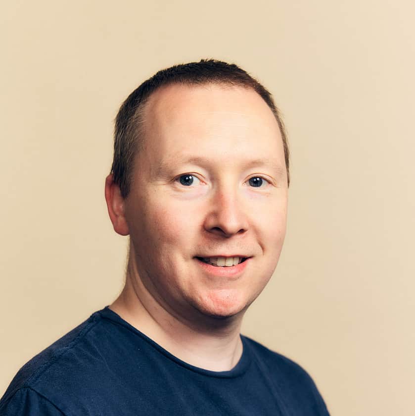 A smiling man with short hair wearing a navy blue T-shirt with an Adelphi logo on the chest.